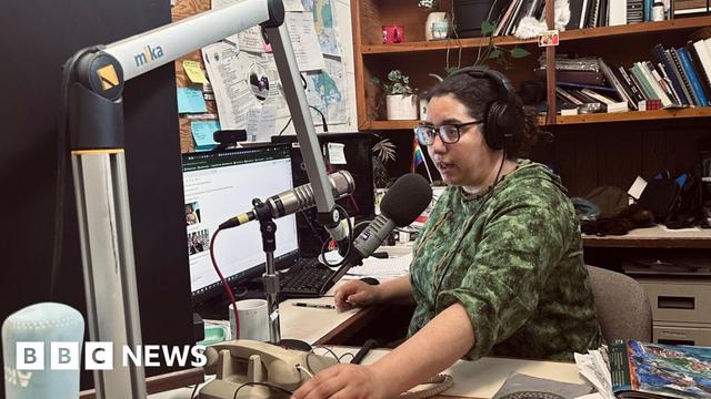 Desire Hagan, wearing a green sweatshirt, glasses and earphones, speaks into a microphone in a radio studio.