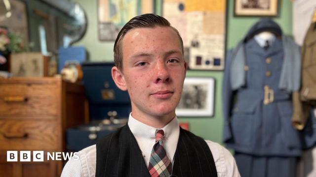 Lincoln smiles at the camera in his bedroom that is decorated in 1940s memorabilia. He has short hair that has been jelled back. He wears a white shirt, tie and black waistcoast. 