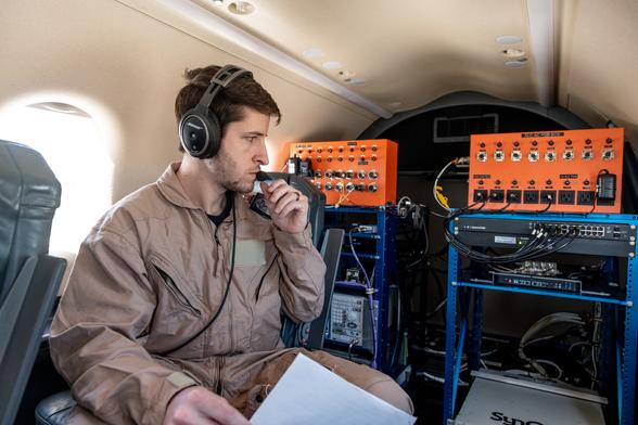 A man in a tan flight suit with a headset and microphone is sitting in a plane holding a piece of white paper. He is looking at communication equipment in the back of the plane. The equipment is comprised of large orange metal boxes with dials connected by wires to a blue computer drive rack. A man in a tan flight suit with a headset and microphone is sitting in a plane holding a piece of white paper. He is looking at communication equipment in the back of the plane. The equipment is comprised of large orange metal boxes with dials connected by wires to a blue computer drive rack.