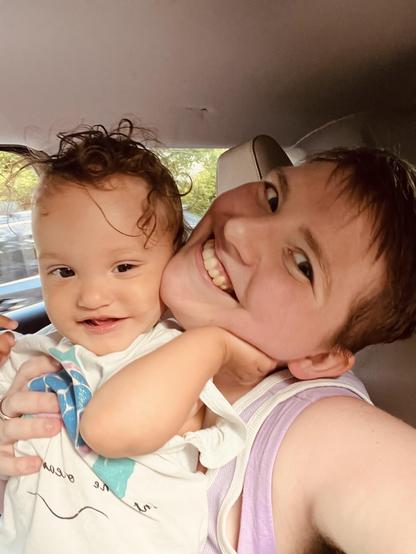 A light-skinned biracial baby with dark curly hair smiles slightly and holds their hand up to the side of their parent's face. The parent, who is white, has their head tilted to the side with a big smile, and has short light brown hair. The background is the inside of a vehicle. A light-skinned biracial baby with dark curly hair smiles slightly and holds their hand up to the side of their parent's face. The parent, who is white, has their head tilted to the side with a big smile, and has short light brown hair. The background is the inside of a vehicle.