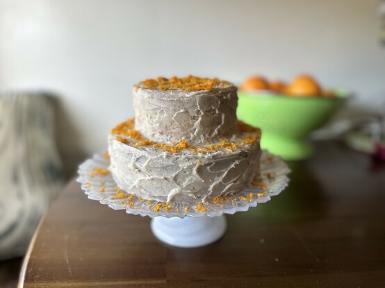 A side view picture of the entire cake. There are two tiers- a four inch cake on top of a six inch cake. It is on a beautiful translucent white cake stand with petal-like edges. The speckled off-white frosting has been applied in a horizontal layered petal pattern, and there is vibrant orange zest sprinkled on top of each layer and the cake stand. In soft focus in the background, there is a bright green bowl of oranges.