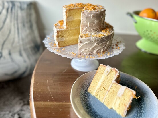 The cake has been cut! The same green bowl of oranges is in the background. But now there is a sliver of cake on a blue plate in the foreground of the photo, in front of the cake. It reveals the layers in the slice and the remaining cake- alternating light orange cake with pale yellow pastry cream.