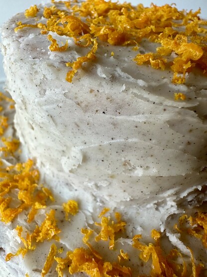 An extreme closeup of the frosting and orange zest. The main focus is on the smooth texture of the frosting and the varying sizes of red-brown cardamom flecks within it, and the way the light is bouncing off the many tiny curled bits of orange peel. 
