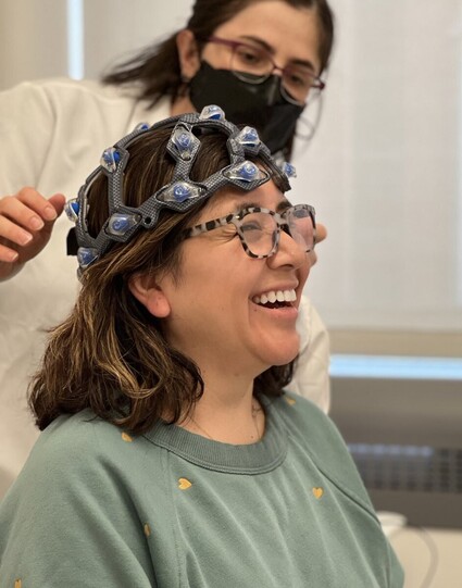 Woman is seated with her right profile facing camera. She is smiling and wearing a green sweatshirt with small yellow hearts. She has short brown hair and glasses. A nurse is blurred in the background with dark hair, glasses, a white lab coat and black mask. She is helping place the WAVi helmet on the participant’s head during her visit to the Center for Clinical and Translational Research (CCTR) our MIT MAESTRO Study!