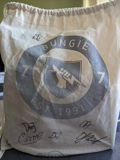 Signed Bungie drawstring backpack in beige, with Bungie est 1991 around the hand-holding-lightning-bolt decal. Signed by members of the team including Cozmo and Deej