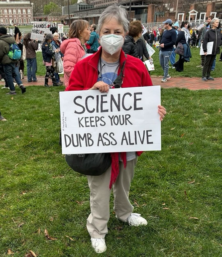 Protestor wearing mask and holding sign that reads “SCIENCE KEEPS YOUR DUMB ASS ALIVE”