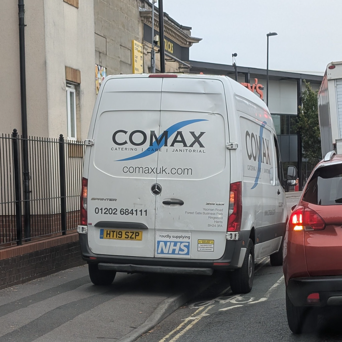 White van marked "Comax", phone number 01202 684111, is parked fully across a pavement at a busy road junction leavinf only enough room for an able-bodied slim person to squeeze past, outer wheels parked on the road edge of a cycle lane, blocking sight of traffic lights.