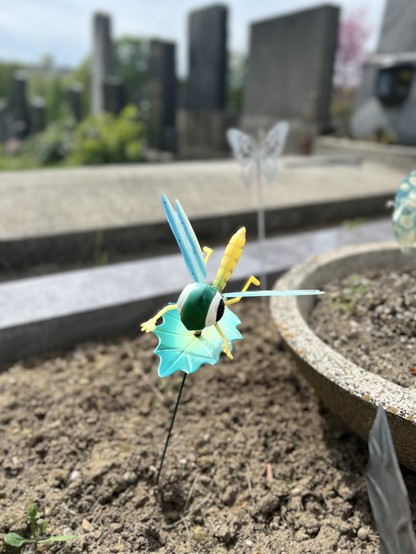 Eine künstlerische Darstellung einer Libelle aus bunten Plastikelementen, montiert auf einem Metallstab im Boden eines Friedhofs, mit unscharfem Hintergrund aus Grabsteinen.
