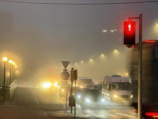 Dichter Nebel am frühen Morgen in der Stadt; auf einer vielbefahrenen Straße stehen Autos und weiße Lieferwagen mit eingeschaltetem Abblendlicht im dichten Verkehr. Eine rote Ampel für Fußgänger ist deutlich sichtbar im Vordergrund rechts. Links führt ein beleuchteter Gehweg mit runden Straßenlampen leicht bergauf. Die Sicht ist stark eingeschränkt durch den Nebel, der die Straßenbeleuchtung diffus erscheinen lässt.