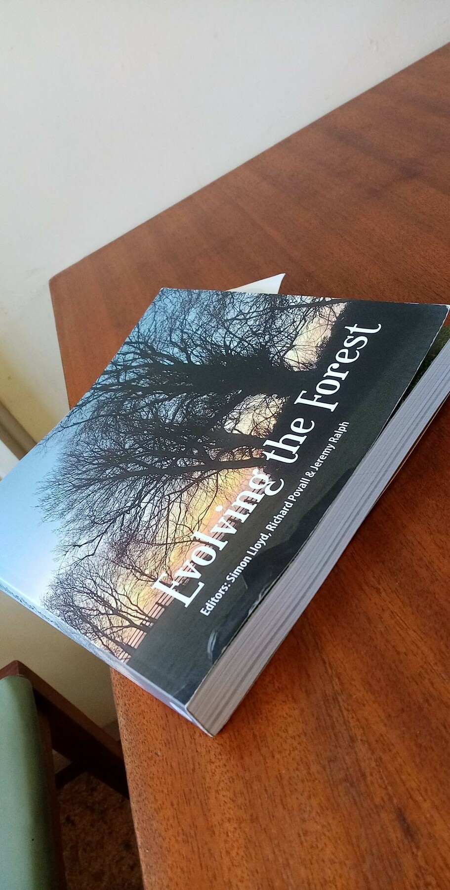 A photograph of a closed book on a wooden table. The cover has a silhouetted photograph of a row of trees. It is called 'Evolving the Forest'.