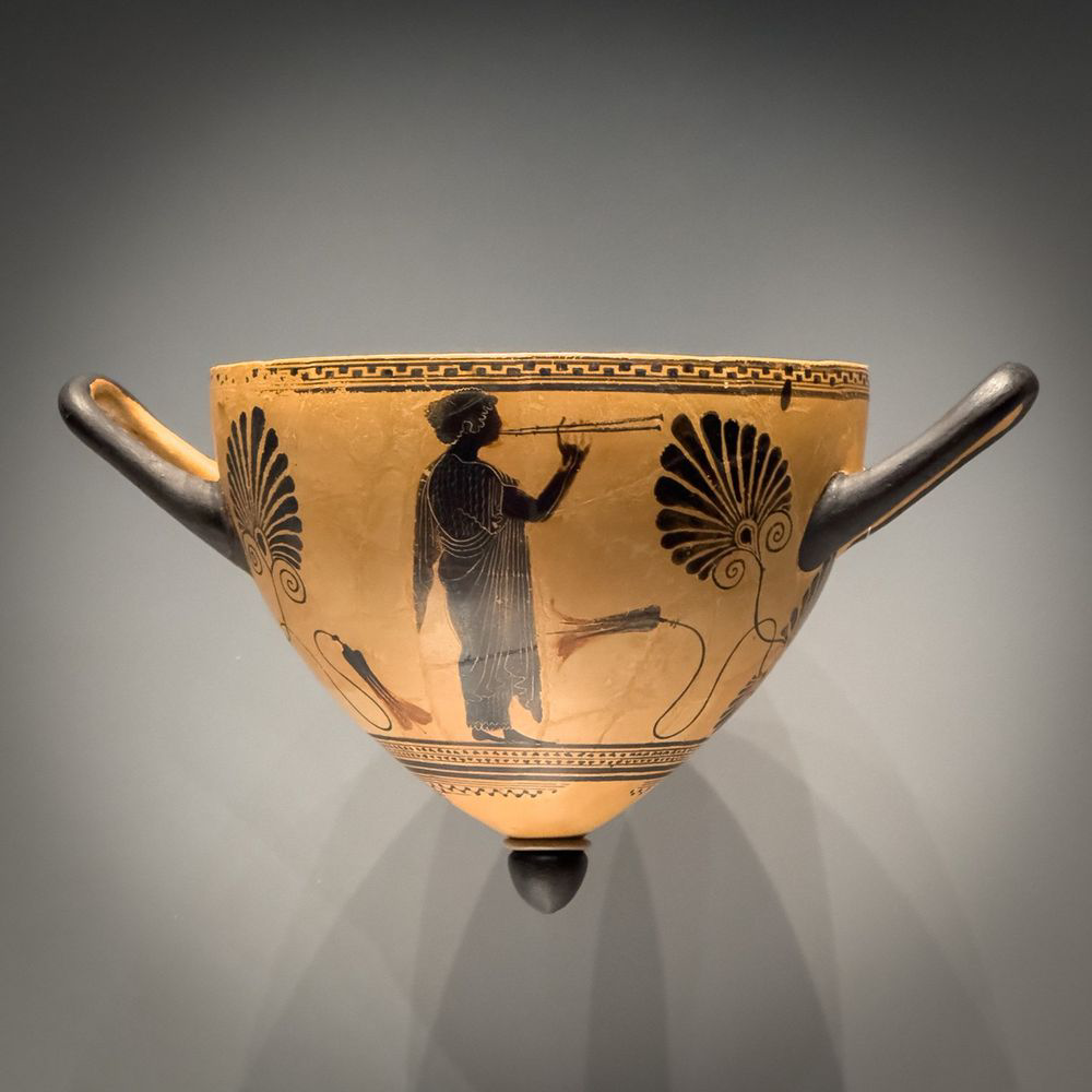 A young woman plays the double flutes on this black-figure mastos or breast-shaped cup. The other side of the vase depicts a woman flourishing a branch and an ivy sprig. These attributes, as well as the nebris or animal skin that she wears over one <br />shoulder, identify her as a maenad, a female follower of Dionysos, the god of wine. A mastos was a drinking cup designed for use at a symposium. A relatively rare form produced by Athenian potters only in the later 500s B.C., it provided a challenge to <br />the drinker. The nipple at the base would not have provided a stable support, and any wine within would have to be consumed before the vessel was put down. Attributed to Psiax, Attic Greek, active 525-510 BCE. Getty Villa Museum. Photo: my own.