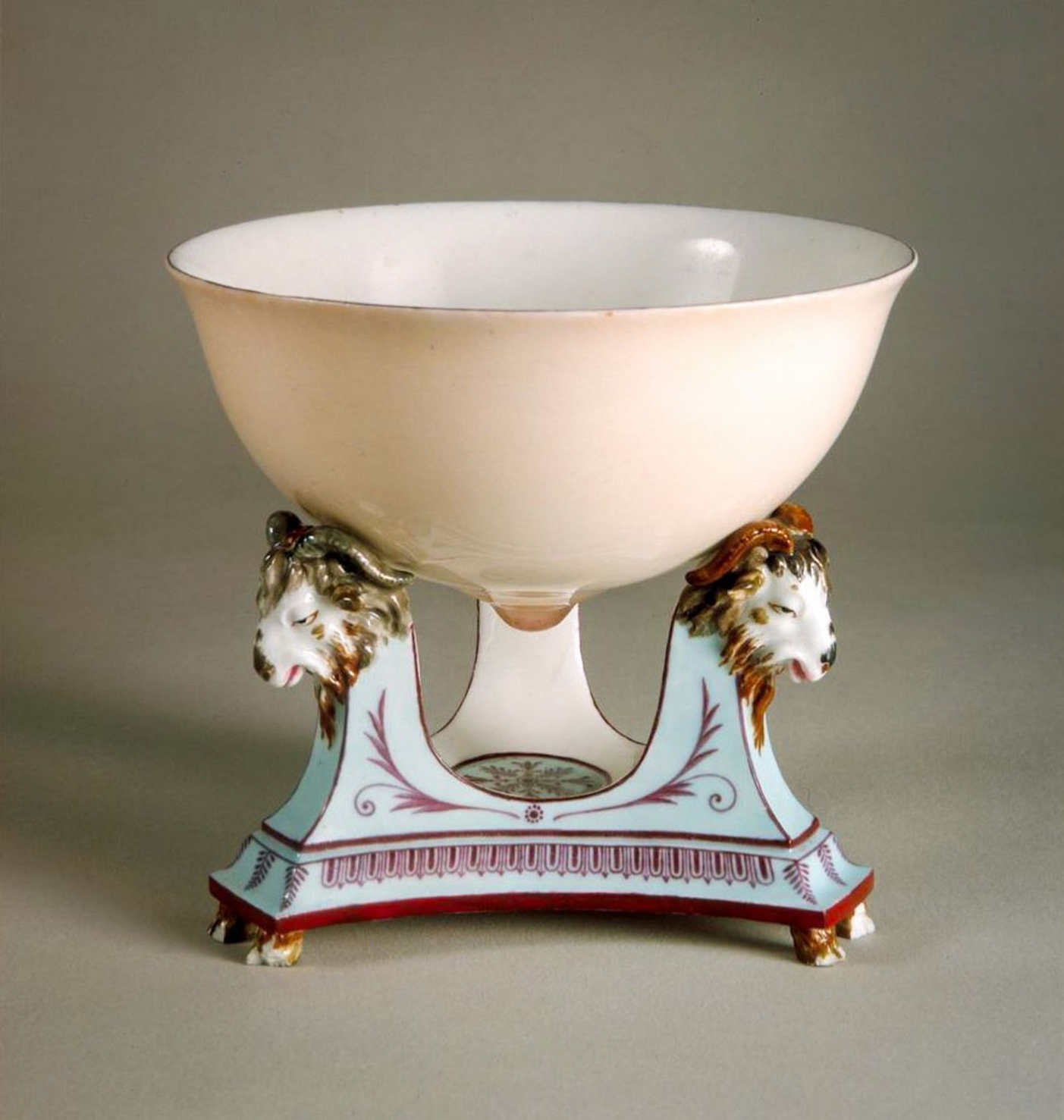 Delicate Sévres porcelain bowl, in the shape of a breast, supported by a porcelain tripod with painted and gilded goat heads. The bowl is a very pale shell pink, with the blushed nipple slightly darker. The tripod below the goats' heads is a pale blue, <br />with dark maroon decorative patterns (somewhat Greek looking). Pairs of little goats' hooves support each leg of the tripod. (Sèvres Porcelain Manufactory, 1788). Painted by Fumez, after a design by Jean-Jacques Lagrené and Louis-Simon Boizot. Photo: M. <br />Beck-Coppola (Musée National de la Céramique).
