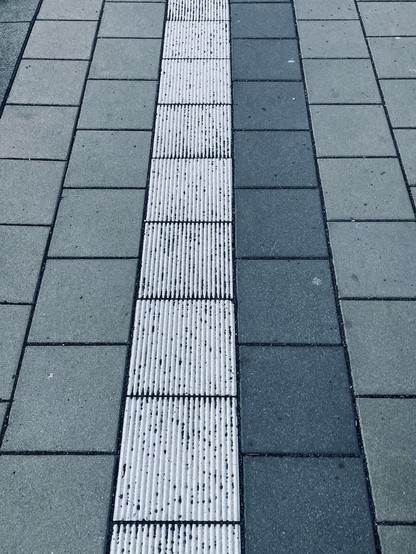 Monochrome photo of a train platform with square stone tiles. In the center, a section of grooved white tiles has accumulated tiny dark pebbles. Sometimes single stones, sometimes many in a row.