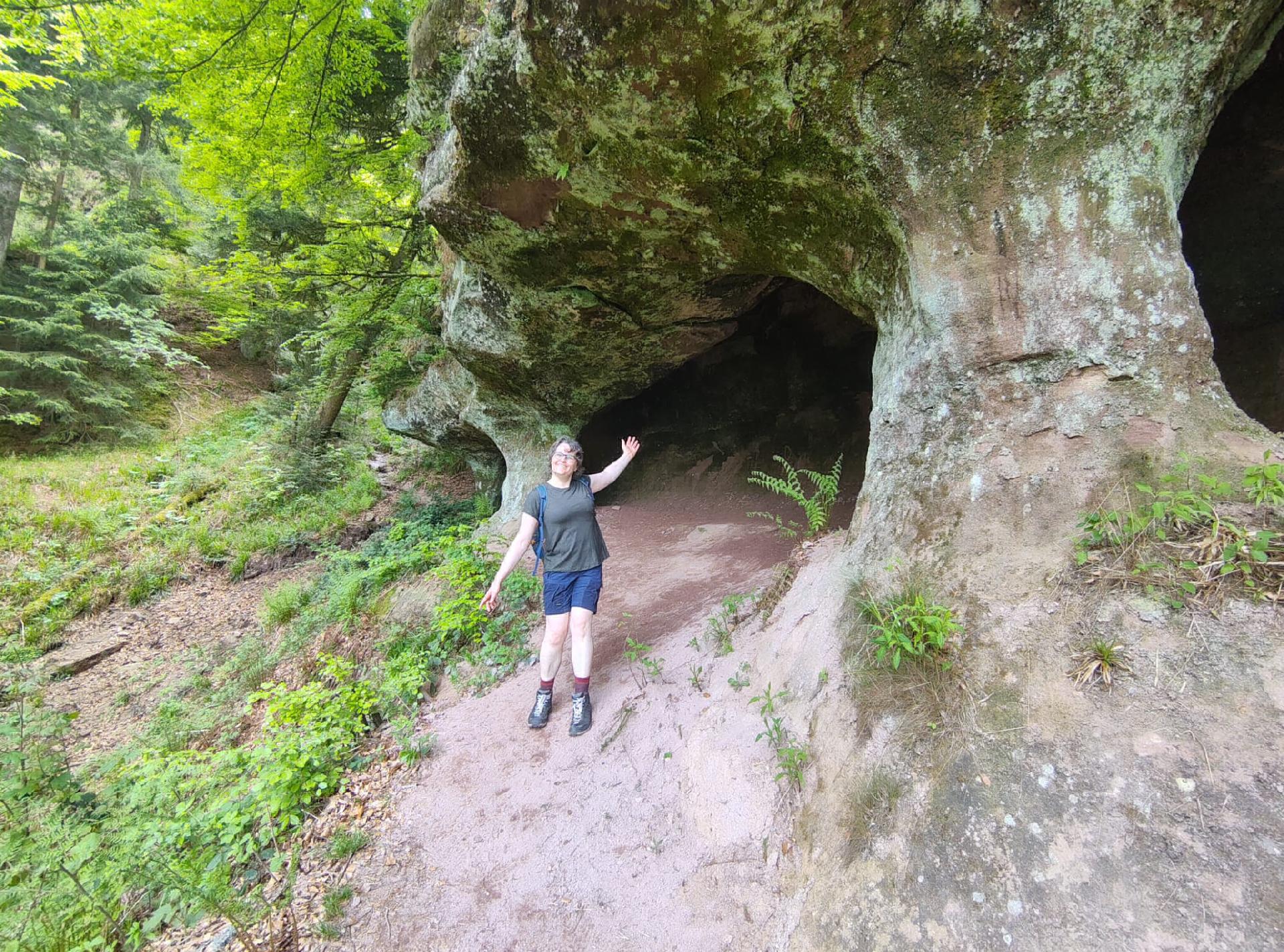 A woman stands in the woods pointing out a large crevice/hole in stone
