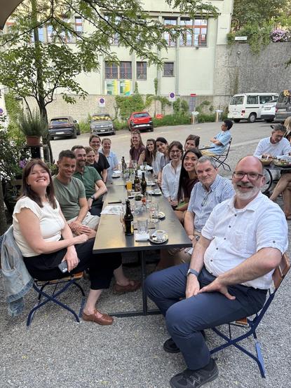 Bunch of smiling people around a table under a tree in a garden restaurant. Drinks pretty much empty and a parking lot in the background.