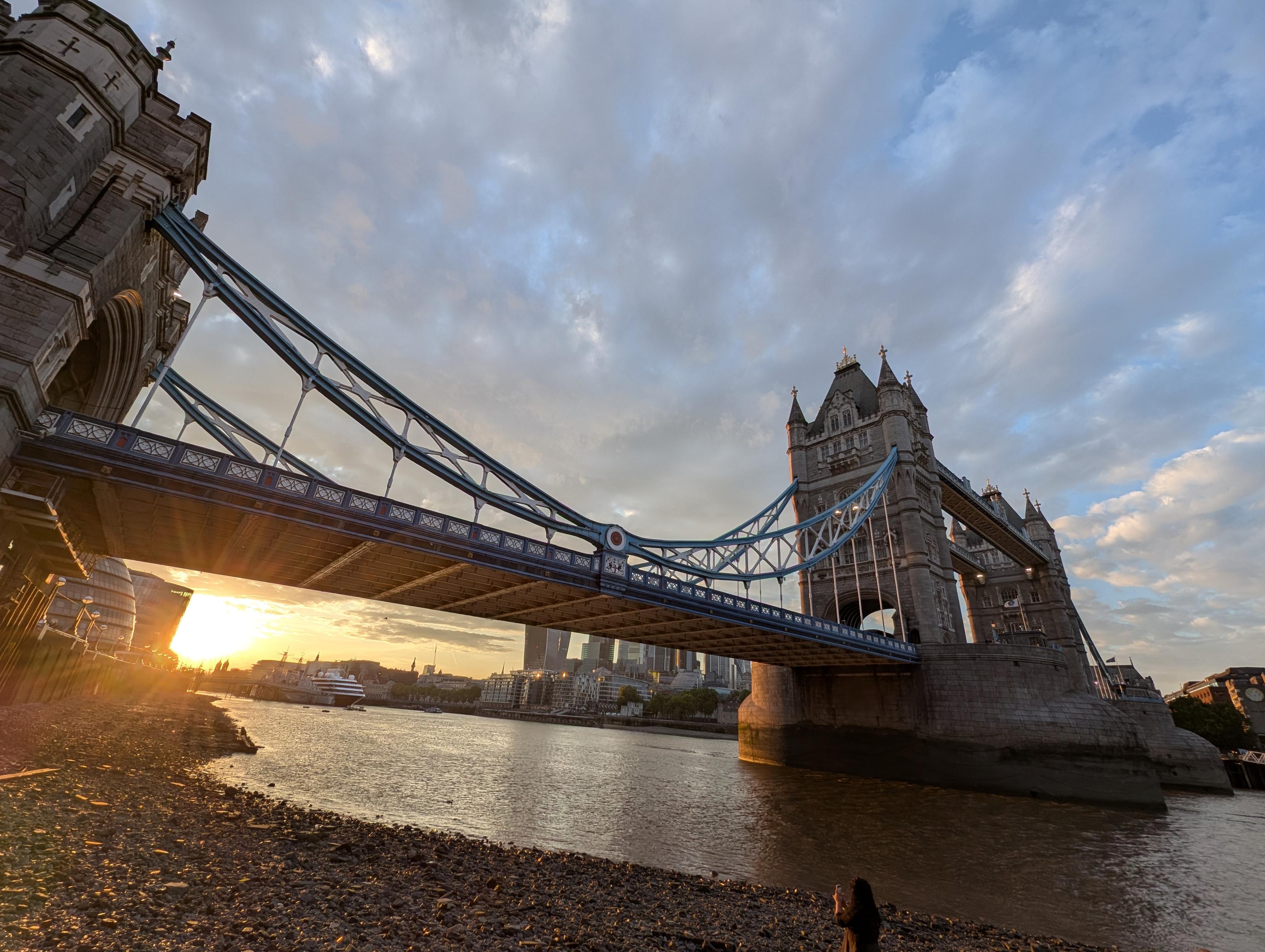 Wide angle view of Tower Bridge in London. From below at low tide, Rocky Beach, underside of bridge visible, sunset in bottom left, reflecting off rocks. Cityscape in distance.