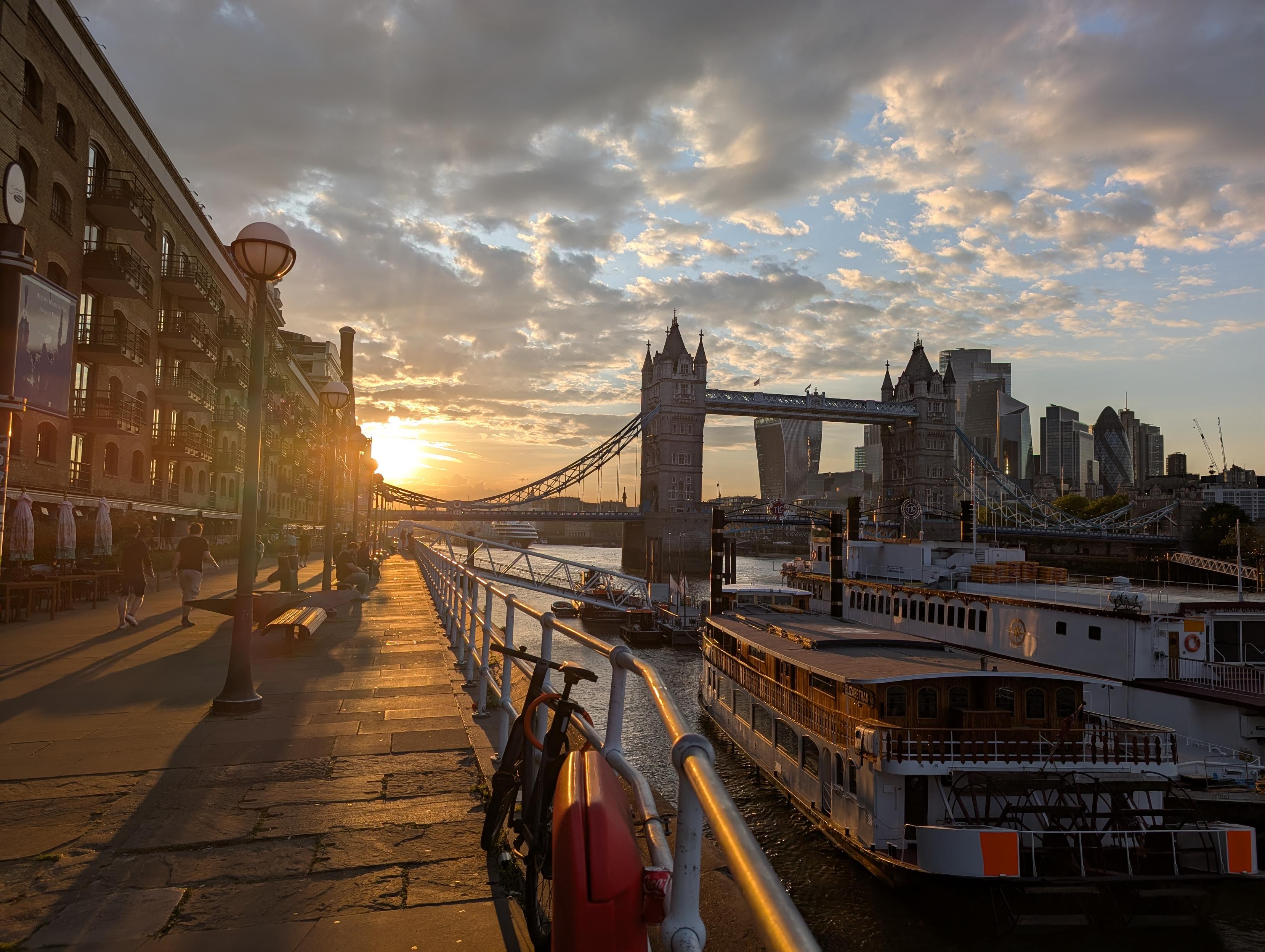 Shot up walkway heading towards Tower Bridge. Boats and barges in water in front of bridge. Sunset on left reflecting off stonework of walkway. London skyline visible in background.