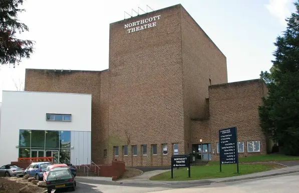 The image shows a building identified as the Northcott Theatre. It has a modern architectural style, featuring a mix of brick and glass elements. The theatre includes signage with its name prominently displayed, and there are cars parked in front of it. Surrounding the building are trees and landscaping, typical of a theater or cultural venue.

Image Credits: Wikimedia / Smalljim / CC BY-SA 3.0
