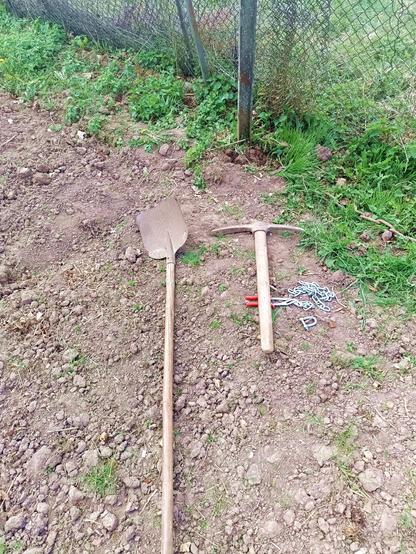 A shovel and a pickaxe laying on the earth. Also some pliers a chain and a shackle. There's some weeds and part of a fence behind.