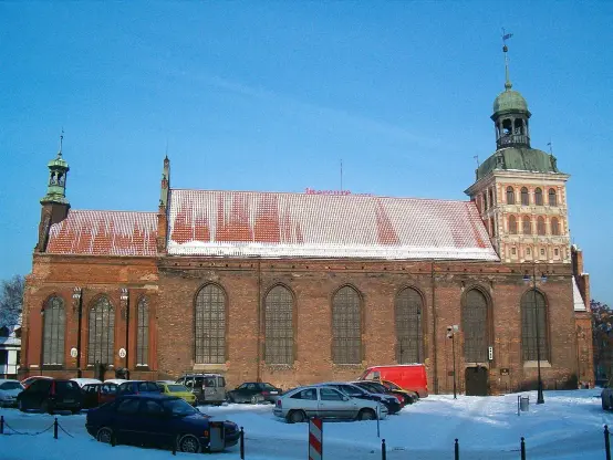 The image depicts a large brick building with a distinctive architectural style, likely a historical or religious structure. It features a red tiled roof and has towers on either side. The surrounding area is covered in snow, suggesting it’s winter. There are several cars parked in front of the building. The sky is clear and blue, adding to the winter scenery.

Image Credits: Wikimedia / Brosen / CC BY 2.5