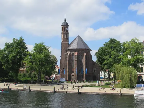 The image depicts a historical building beside a body of water, likely a river or lake. The structure features a distinctive architectural style with a conical roof and a tower. Surrounding the building are trees and a walkway where people are seen walking and cycling. The scene appears to be in a pleasant urban or semi-urban environment, providing recreational space for visitors.

Image Credits: Wikimedia / Gregor Rom / CC BY-SA 4.0