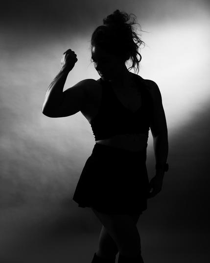 Silhouette of a person flexing their arm against a gradient background. Hair is tied up, and the person wears a sleeveless top and skirt. Contrasting light emphasizes the muscle and form.