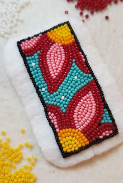 an in progress beaded panel on felt by kiamichi depicting a large fraction of two flowers with red and pink petals with gold and yellow centers in the top right and bottom left corners of a panel on a teal background with a black beaded frame. surrounding the panel of felt are piles of beads in red, white, and yellow