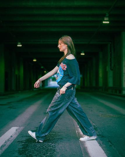 A person wearing a dark graphic T-shirt and baggy jeans walks across a street in a dimly lit tunnel. The scene features overhead lights casting a greenish hue, and the person wears white sneakers and bracelets on their right wrist. The image captures motion with the person's arm slightly extended.