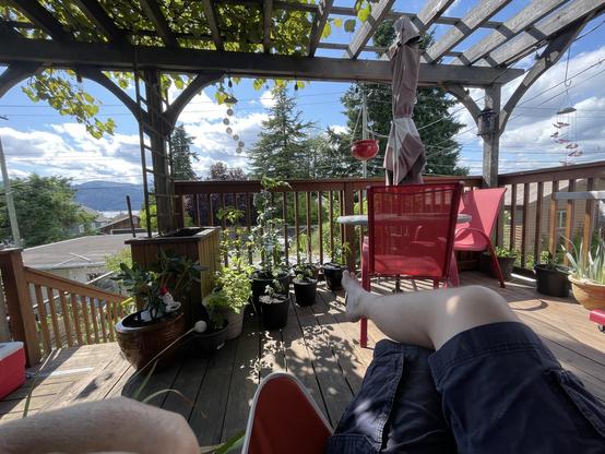 My legs jut out crossed in front of me aa I sit on the deck. There are a number of plants in pots in front of me beside the deck railing. There are two red chairs around a small table to the right. To the left is the stairs going down toward the yard. In the distance over the stairs the Inlet can be seen under a blue sky and partly cloudy