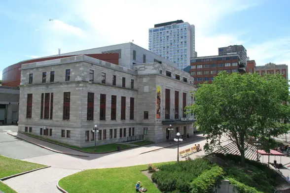 The image features a building with a modern design, likely a cultural or civic structure, surrounded by green space and pathways. It appears to be a sunny day, and there are trees and benches visible in the vicinity. The overall setting suggests a public area, possibly in an urban environment.

Image Credits: Wikimedia / Malimage / CC BY-SA 3.0