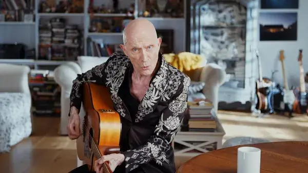 A still from the film Strange Journey: The Rocky Horror Story. A thin, bald white person with notably pointy ears, wearing an elaborately patterned black and white jacket, sits in a bright living room playing an acoustic guitar.