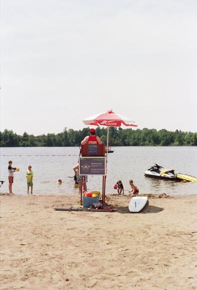 Lifeguard sitting under a red umbrella on a lake beach in Ottawa, Canada. Children are playing. The lifeguard is in a bright red outfit.