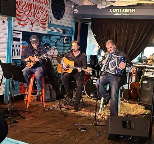 Three musicians perform on stage in a lively venue. One plays a mandolin, another strums an acoustic guitar, and the third plays an accordion. The backdrop features colorful artwork, and various musical instruments are visible around them.