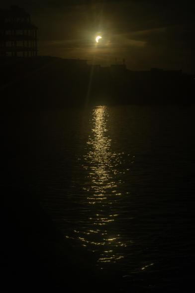 A yellowish sun with a bite out of it to the bottom right as the moon passes above it. Below a long smear of golden reflections ripples across the surface of a river towards the viewer.