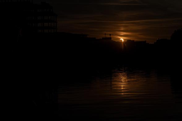 The sun twinkles as it kisses the horizon. The bottom left of it is obscured by the moon and it turns the sky and gently rippling river a rich bronze.