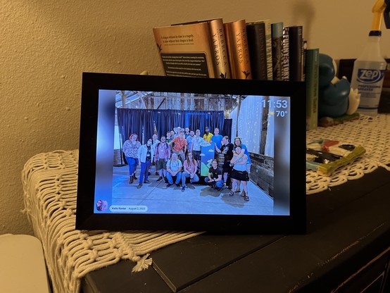 Digital photo frame on a dresser by some books, featuring a group photo from North Bay Python 2023