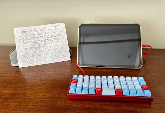 On a hotel room desk sits a yellow Apple iPad in a pink folio case, propped up in landscape orientation. Connected to it is a blue and red OLKB Planck keyboard, and to the left is a printout of the keyboard layout.