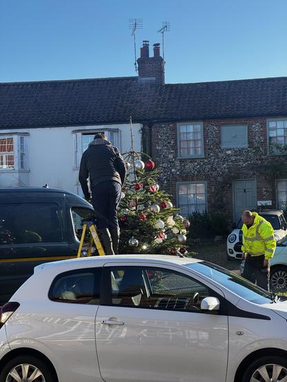 Two guys, one on a ladder and one watching from the side, putting decorations on a Christmas tree, on the green in the village. There’s a car in the foreground and Terence houses behind