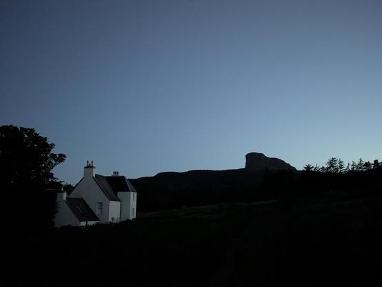 after sundown, to the left the white walls of an old manse still visible in the evening glow, to the right the dark silhouette of a hill with a distant peak with vertical cliffs