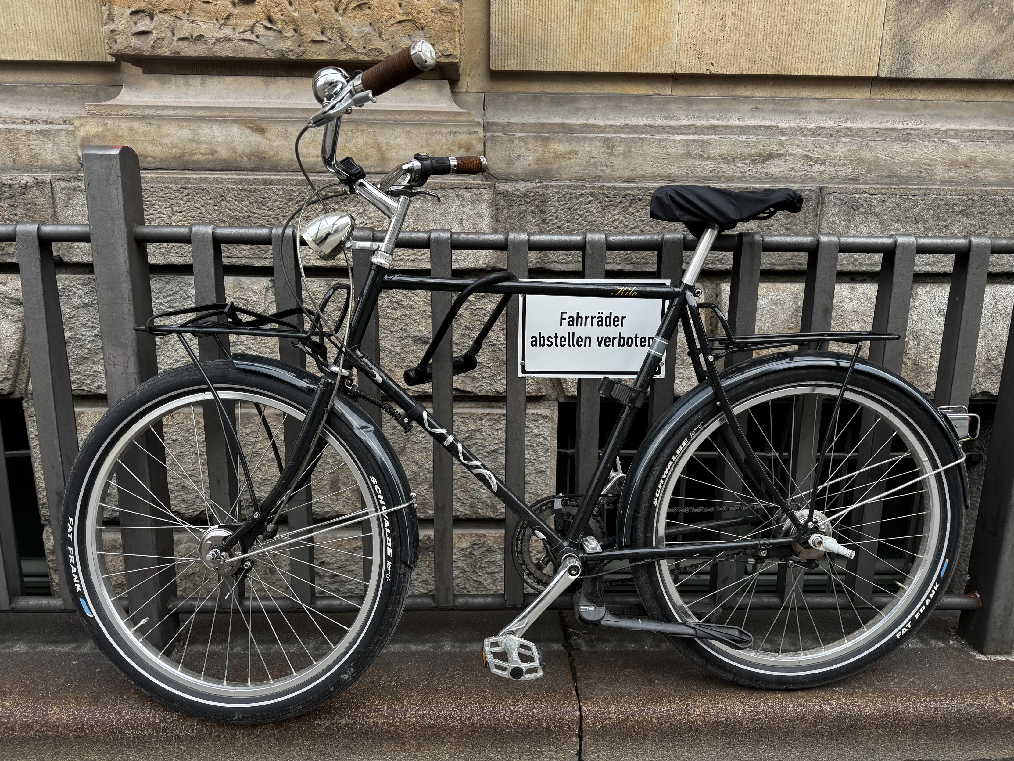 Ein Fahrrad mit großem Lenker und Gepäckträgern vorne und hinten wurde an einem Zaun abgestellt. Am Zaun hängt mittig vom Fahrrad ein Schild auf dem steht „Fahrräder abstellen verboten“.