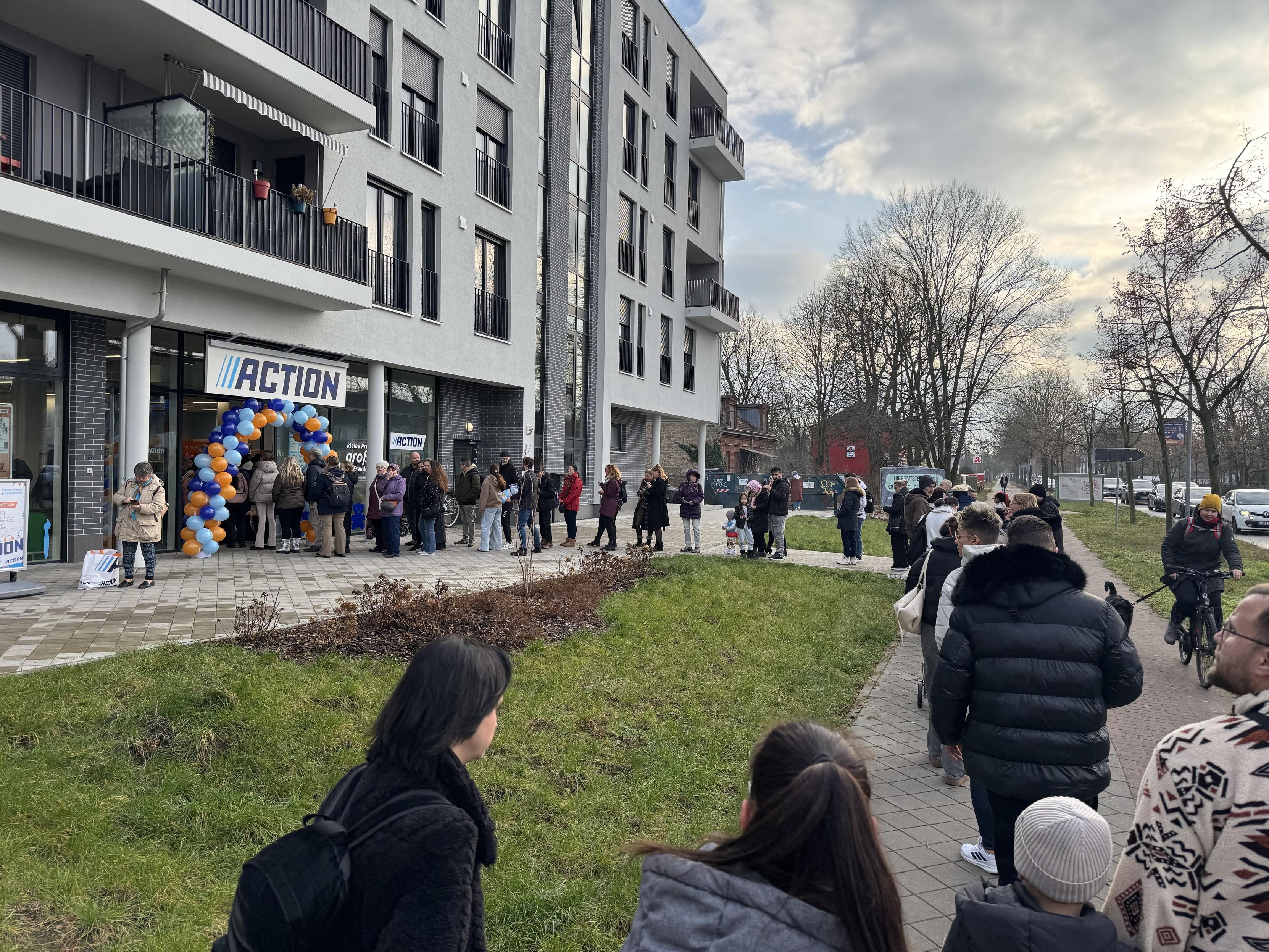 A long line of people waiting outside a store labeled "ACTION," decorated with an arch of blue and orange balloons. The setting includes a modern building with balconies and greenery in the foreground, under a cloudy sky.