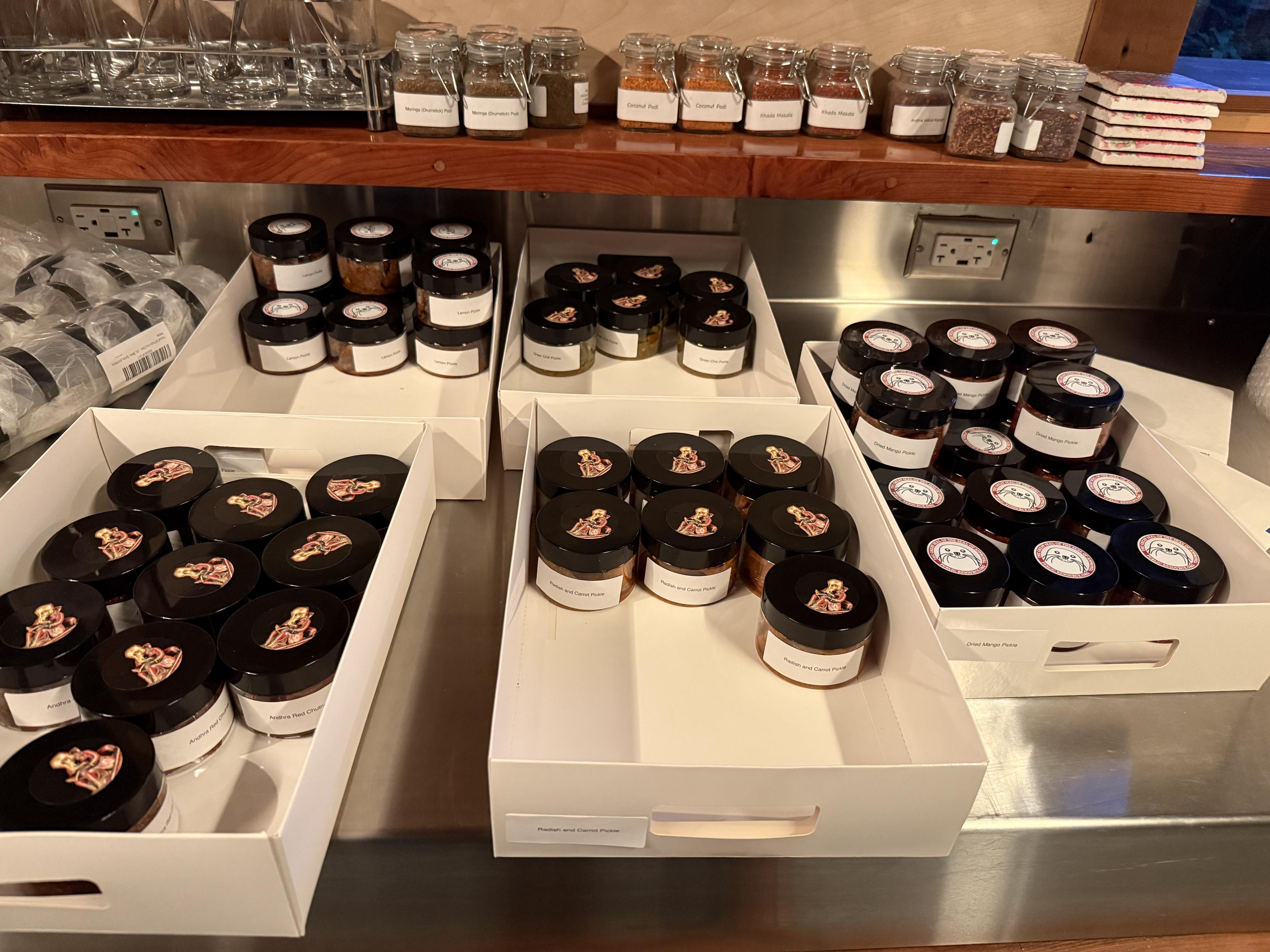 Five flats, each with 12-30 containers of pickles. The jars are 4 ounces each. The shelf over the flats have jars of masala blends. 