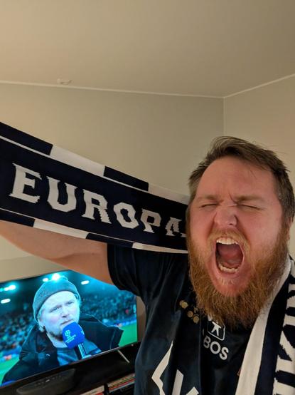 Ein mann med raudbrunt skjegg og knea augo ropar i rein ekstase mot kameraet. Han har på seg ei mørkeblå Viking FK-drakt og held opp eit mørkeblått og kvitt supporterhalstørkle der det står «EUROPA» med store bokstavar. I bakgrunnen syner ein TV-skjerm ei fotballsending med ein mann som snakkar i ein mikrofon.