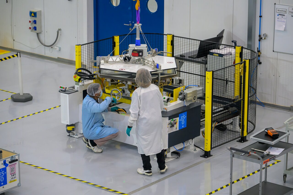A lab with a flat hexagonal object held above a structure by a rope and a complex support. Two people wearing hairnets and lab coats are inspecting it.