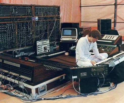 A picture of electronic music pioneer Klaus Schulze seated on the floor surrounded by various synthesizes and other electronic/electric musical equipment. The gear includes a large Moog 55 modular system, a mixer console, a Mini Moog, a Farfisa organ and and VCS3.