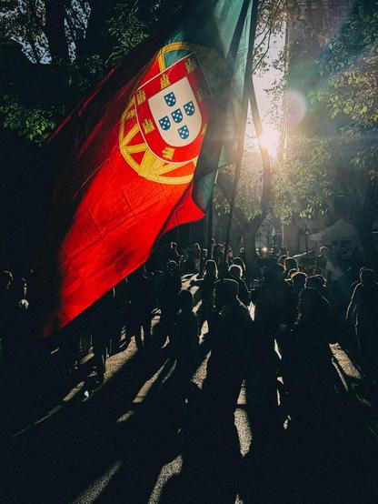 Large Portuguese flag waving above a crowd of silhouetted people on a sunlit street with long shadows and trees in the background.