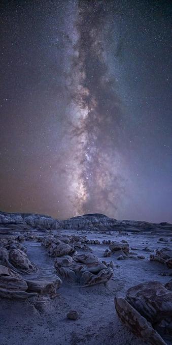 The Milky Way over the Cracked Eggs in New Mexico’s Bisti Wilderness, presented as a tall, narrow panorama.

The cracked eggs are oddly shaped rocks that have reminded people of alien eggs. They are a small, tight grouping, each one the same but also, different. Together, they make an abstractive grouping. We’re standing in the middle of them, and they lead to the distant hills. Each rock looks like an egg with wings, and they are between knee and waist height.

The Milky Way is vertical in the scene, letting me make it narrower than usual. It’s fairly bright, and there are some reddish bands of airglow in the lower left of the sky. In fact, thanks to air glow and the atmosphere, the night sky is colorful, with blues and greens and colors in between. 

The overall tones are dark and subdued, it being night and all.