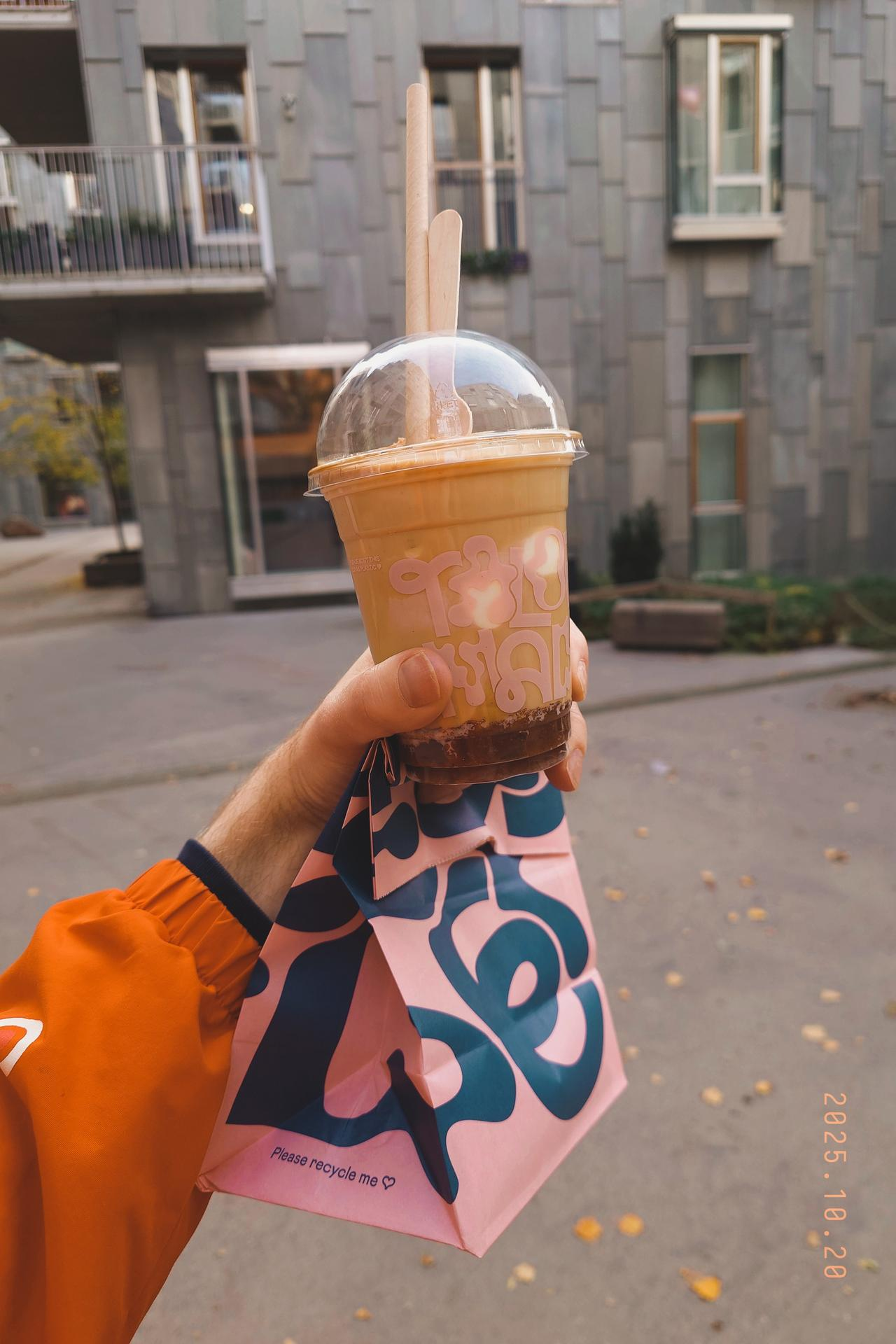 A photo of Matt's hand holding a plastic cup containing a very sweet looking coffee drink - with layers of stewed apple, caramel, milk, coffee, ice cream, crumble, and a spoon and straw sticking out the top. He's also holding a Talormade branded bag containing a donut.