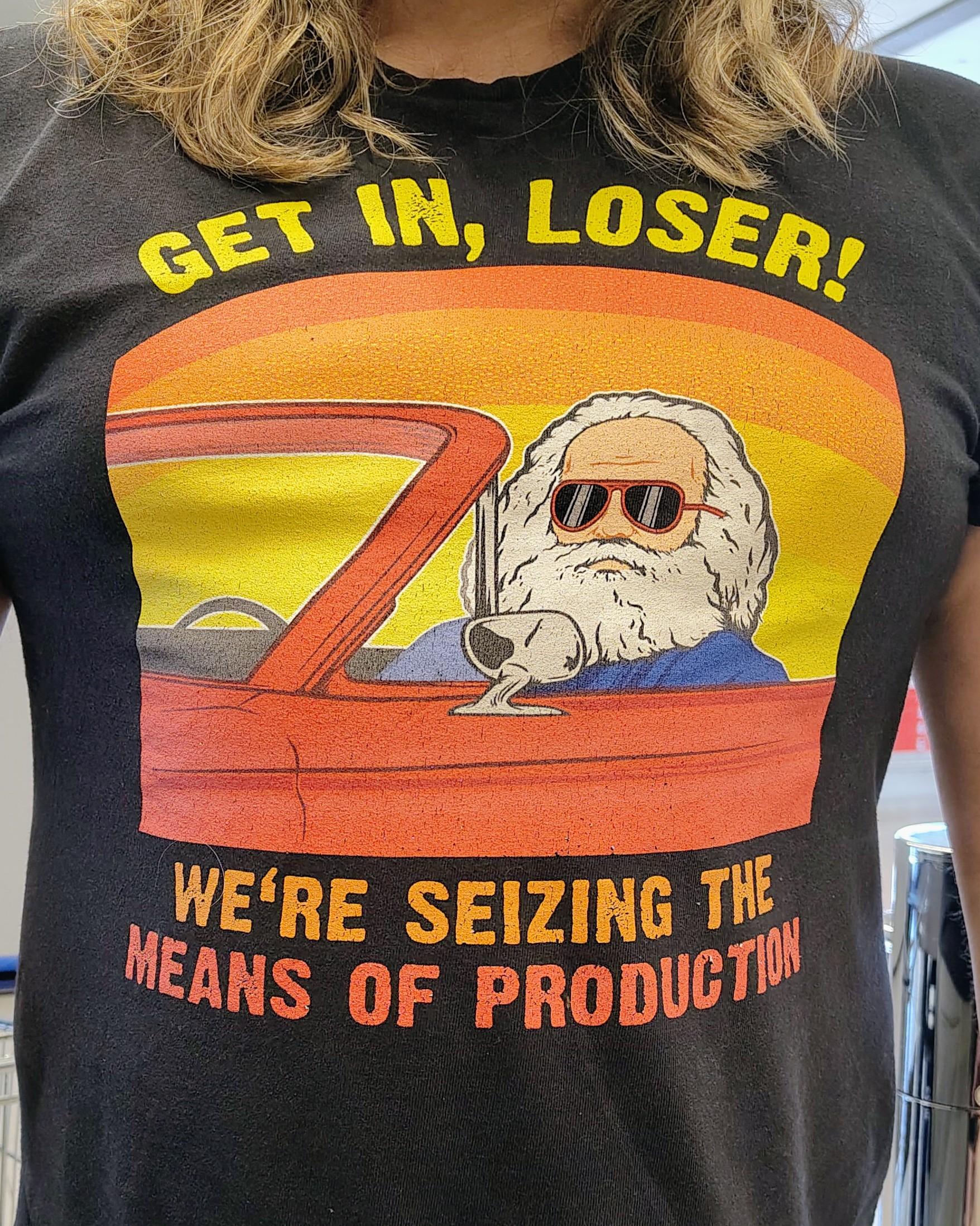 En overkropp i svart t-skjorte med tegning av Karl Marx med solbriller sittende i en rød sportsbil. Over tegninga står det: "Get in, loser!" Under står det: "We're seizing the means of production"