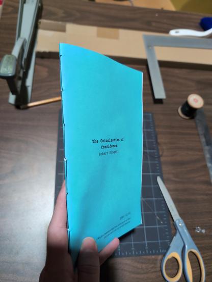 A pamphlet of The Colonization of Confidence by Robert Kingett, with a blue cover, being held so that the black stitched binding can be seen. In the background is a worktable with scissors, a cutting mat, thread measuring tools and the stapler I'd been using.